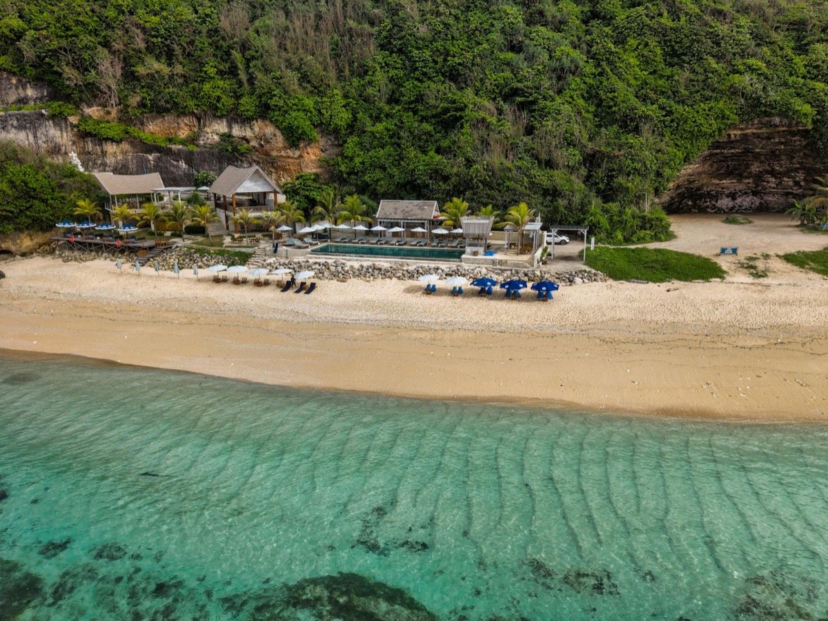 Aerial view of luxury beachfront resort with turquoise water, sandy beach, and lush tropical hillside backdrop